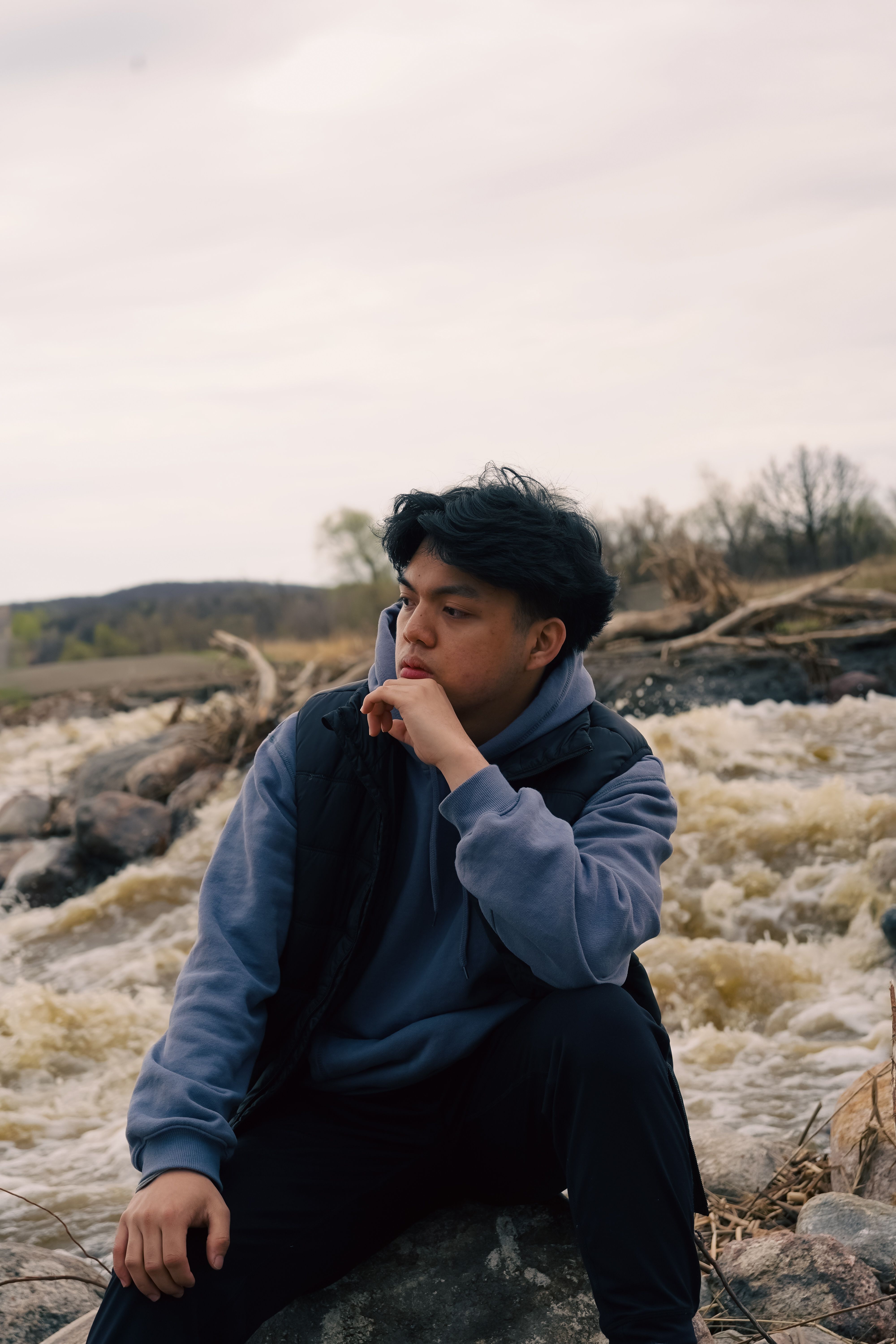 A young man in a blue hoodie sits pensively by a rushing stream outdoors.