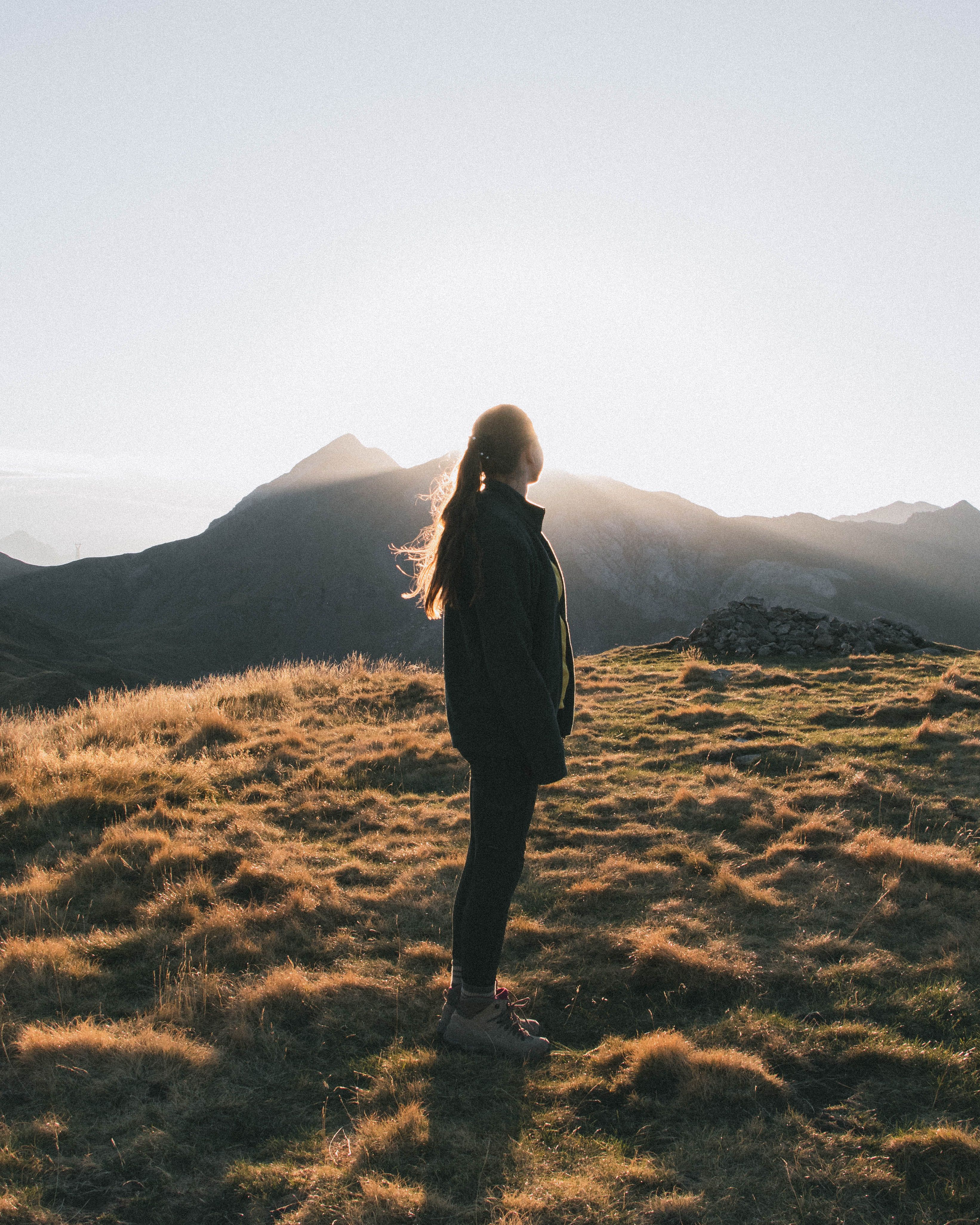 A woman stands silhouetted on a grassy mountain at sunset, capturing a moment of tranquility and natural beauty.