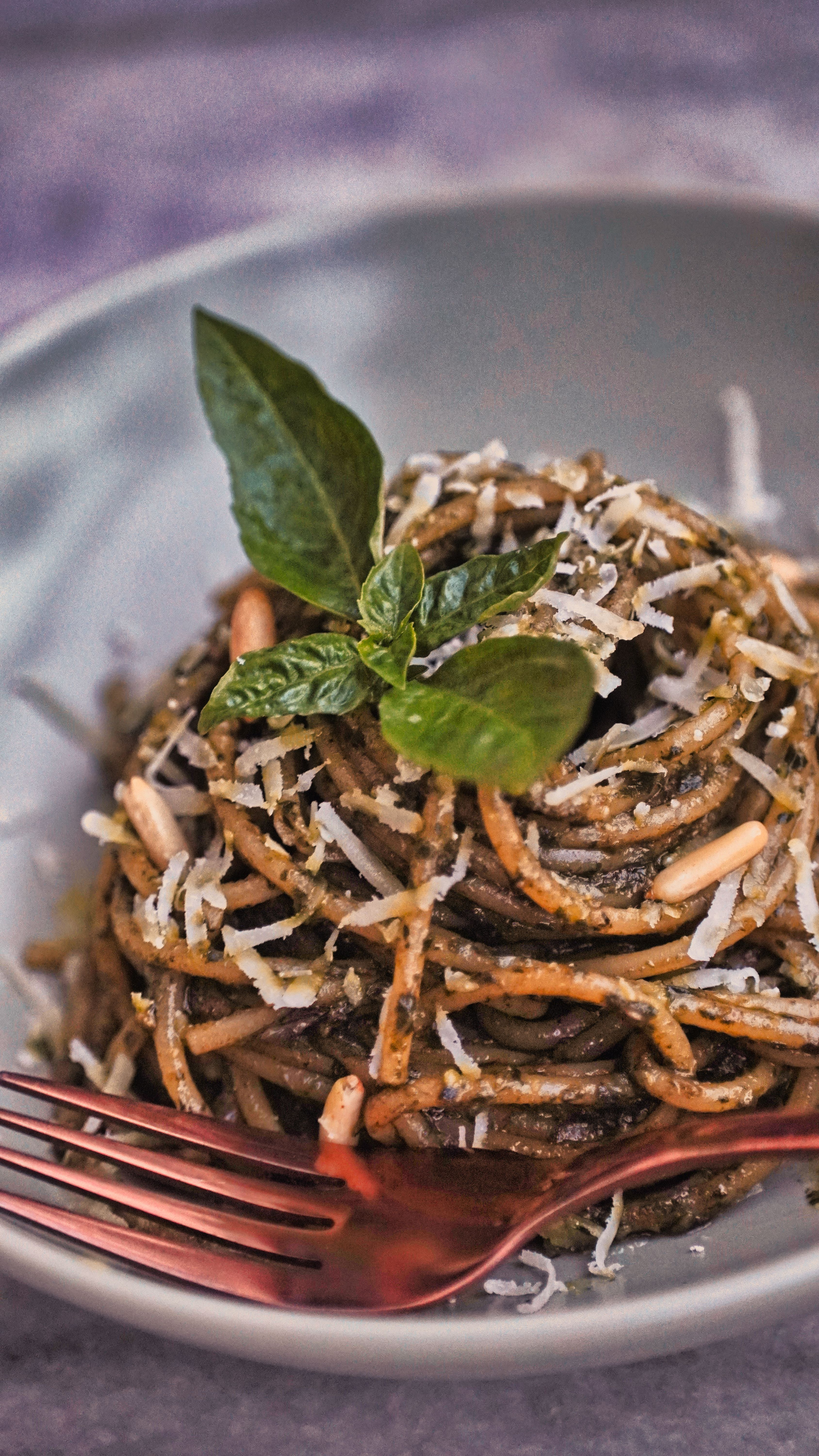 A rustic bowl of bright green basil pesto studded with toasted pine nuts, with a wooden spoon resting inside and loose basil leaves and whole pine nuts scattered on the counter beside it.