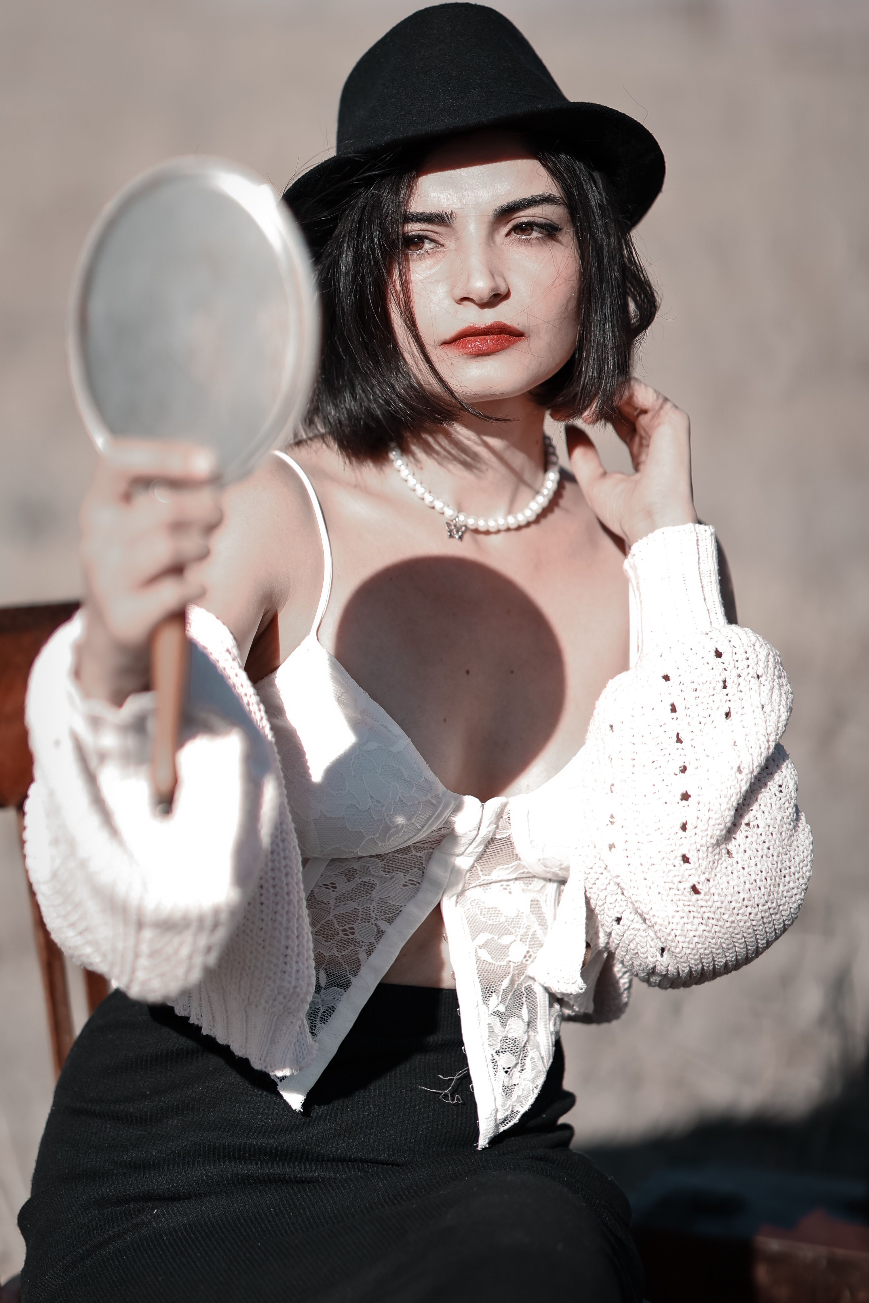 A stylish woman in a hat holds a mirror, reflecting elegance and fashion in outdoor sunlight.