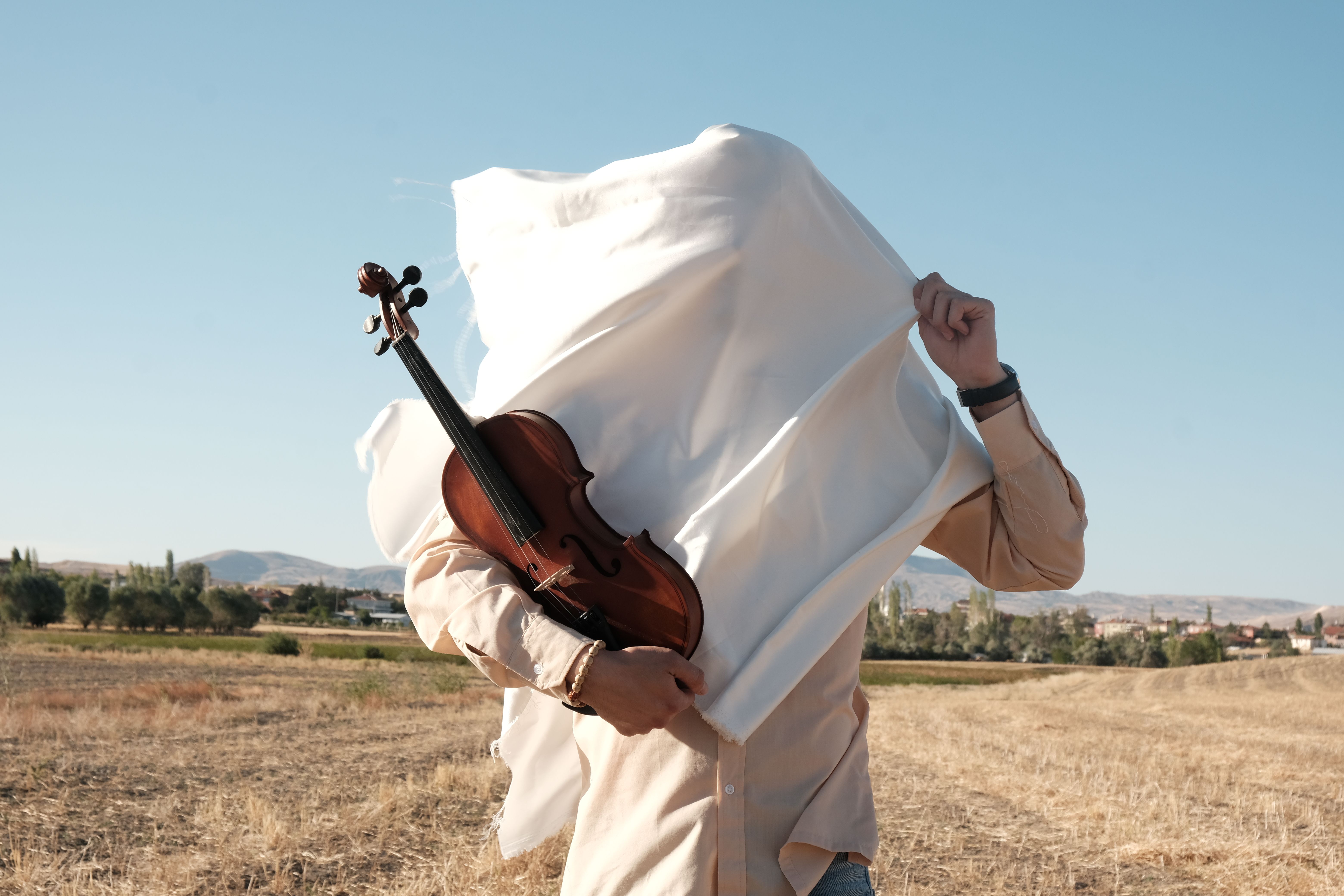 A person holding a violin beneath a flowing cloth in a sunny rural field.