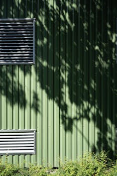 A green corrugated metal wall with shadows of leaves and ventilation grilles in summer.