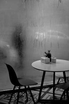 Black and white image of a café table and chairs with a fogged window.