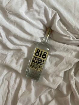 A 818 Tequila Blanco bottle on rumpled white bed sheets, creating an elegant scene.