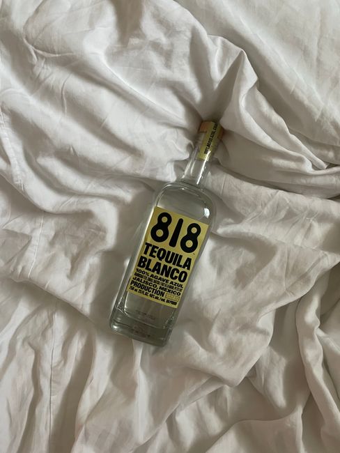 A bottle of blanco tequila next to a rocks glass, lime wedges, and coarse salt on a wooden bar surface