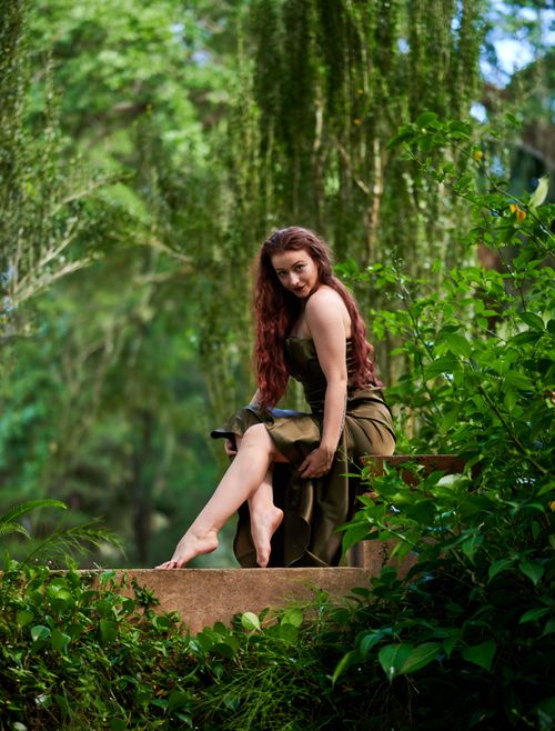 Free A redheaded woman in a green dress poses outdoors surrounded by lush greenery. Stock Photo
