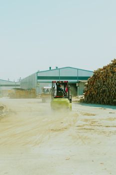 Forklift moving logs in a dusty lumber yard outside a warehouse.