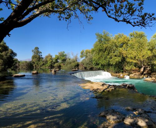 Free Scenic waterfall amidst lush trees and flowing river under clear blue sky. Stock Photo