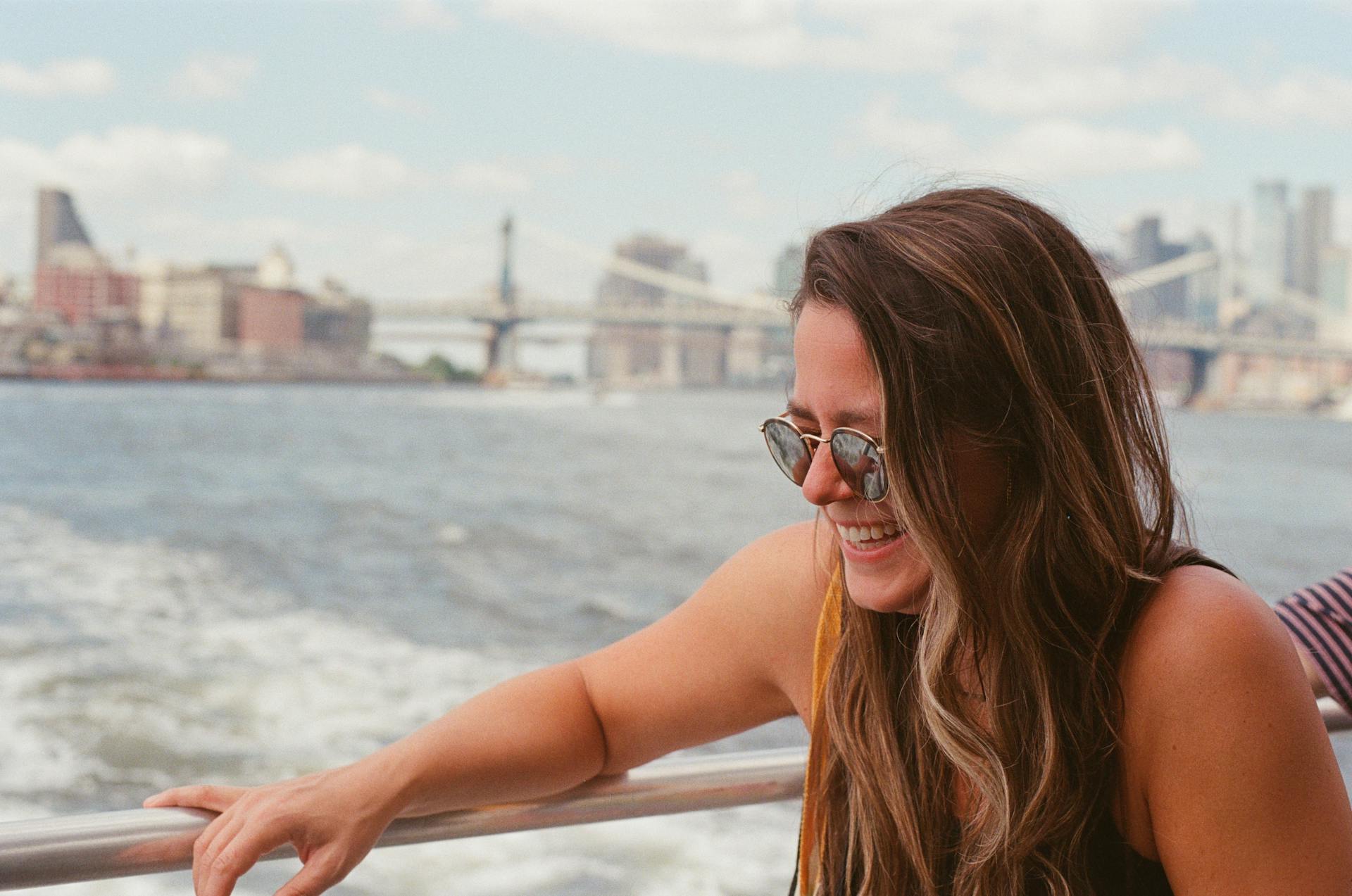 Smiling woman in sunglasses enjoys a summertime boat ride with New York City skyline in the background.