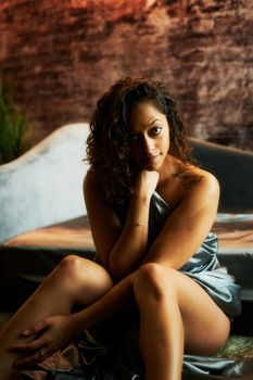 Stylish young woman with curly hair posing on a sofa in a satin dress, exuding elegance and confidence.