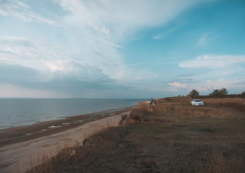 Free A peaceful beachside view featuring a car parked by the coast under a vast blue sky. Stock Photo