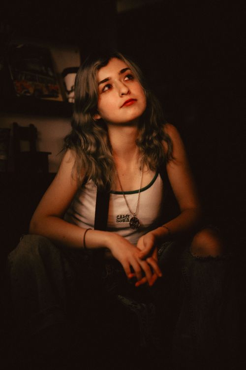 Free A teenager in a tank top sits thoughtfully indoors, surrounded by moody lighting. Stock Photo