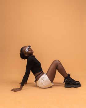 African woman in trendy black top and boots, posing in studio with orange background.