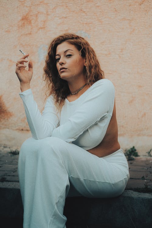 Woman Dressed in White Sitting on Curbside Smoking Cigarette