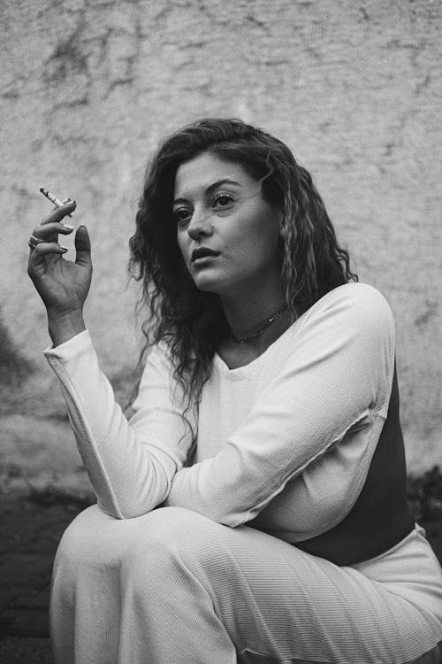 Black and White Photo of Woman Sitting under Wall Smoking Cigarette