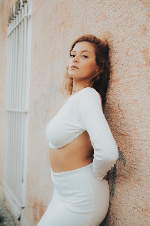 Young Woman in a White Crop Top with Long Sleeves and Skirt Leaning Against the Wall