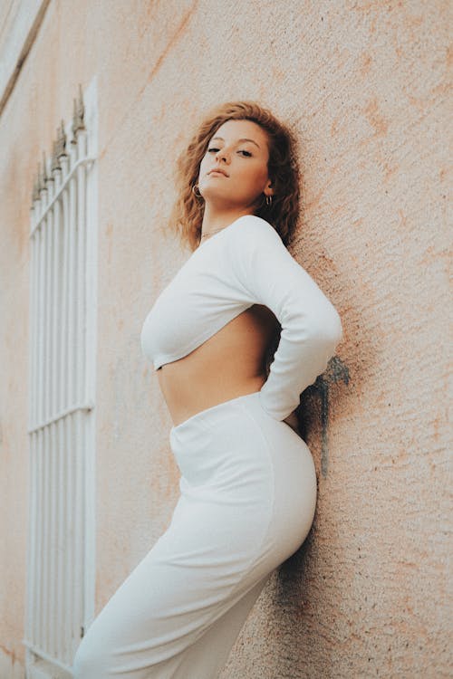 Young Woman in a White Crop Top with Long Sleeves and Maxi Skirt Leaning Against the Wall of a Building