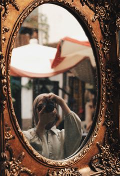 A woman takes a selfie in an intricately designed antique mirror, capturing the vibrant street scene behind her.