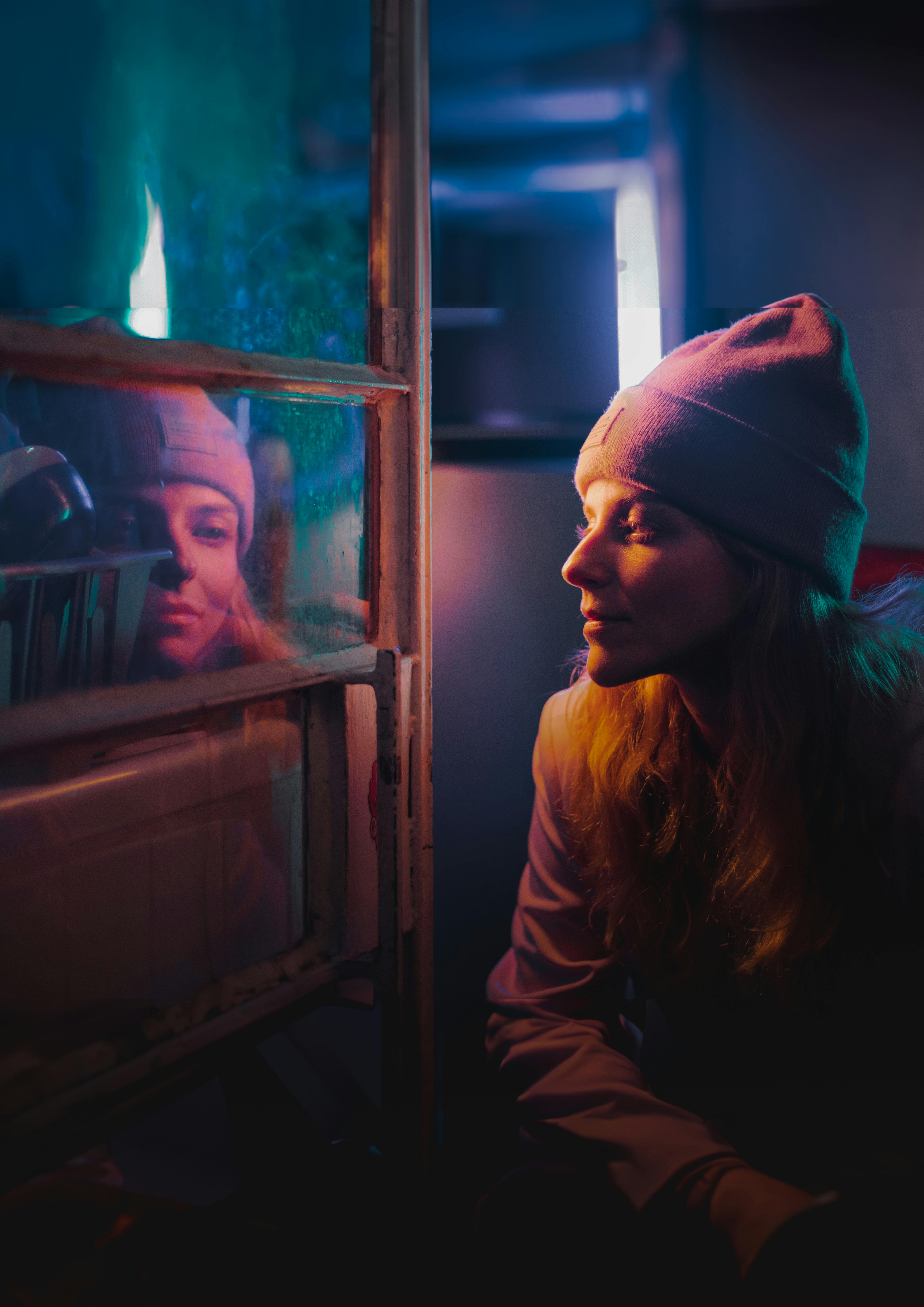 A woman wearing a beanie gazes at her reflection in an illuminated window at night.