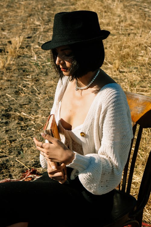 Woman in Black Fedora Hat and White Knit Cardigan Sitting on a Chair in a Field