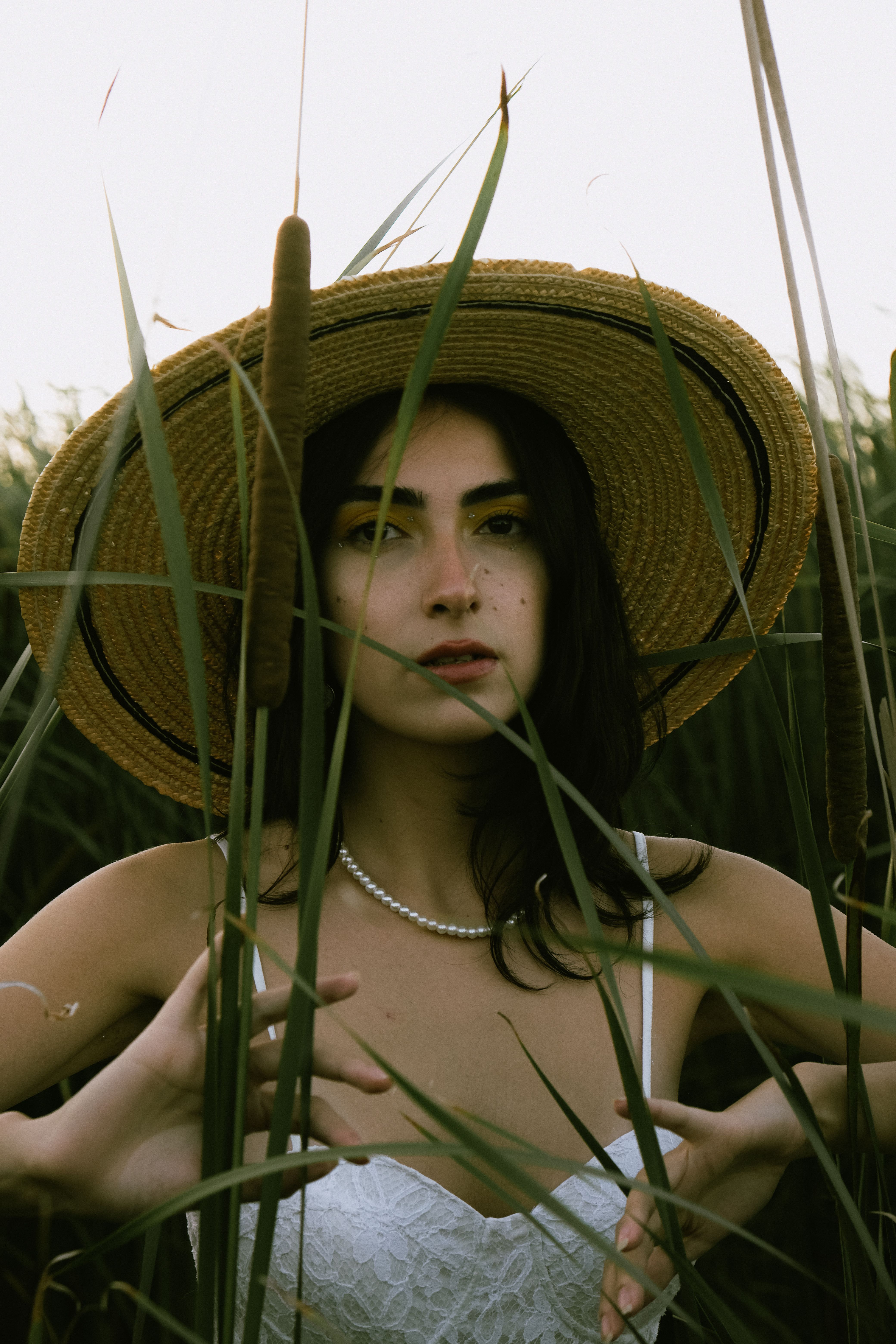 A brunette woman in a straw hat standing among tall reeds in a summer field.