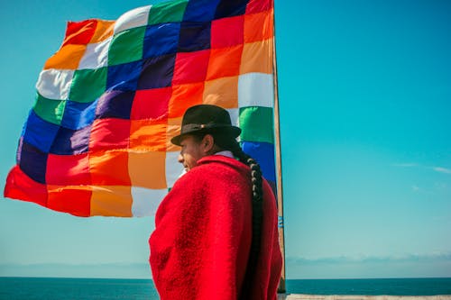 A Man with Braided Hair, Wearing a Hat and a Red Cape Holding a Colorful Flag 