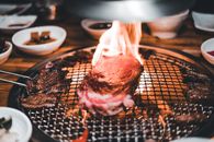 Close-up of a steak grilling with flames on a barbecue, surrounded by dishes.