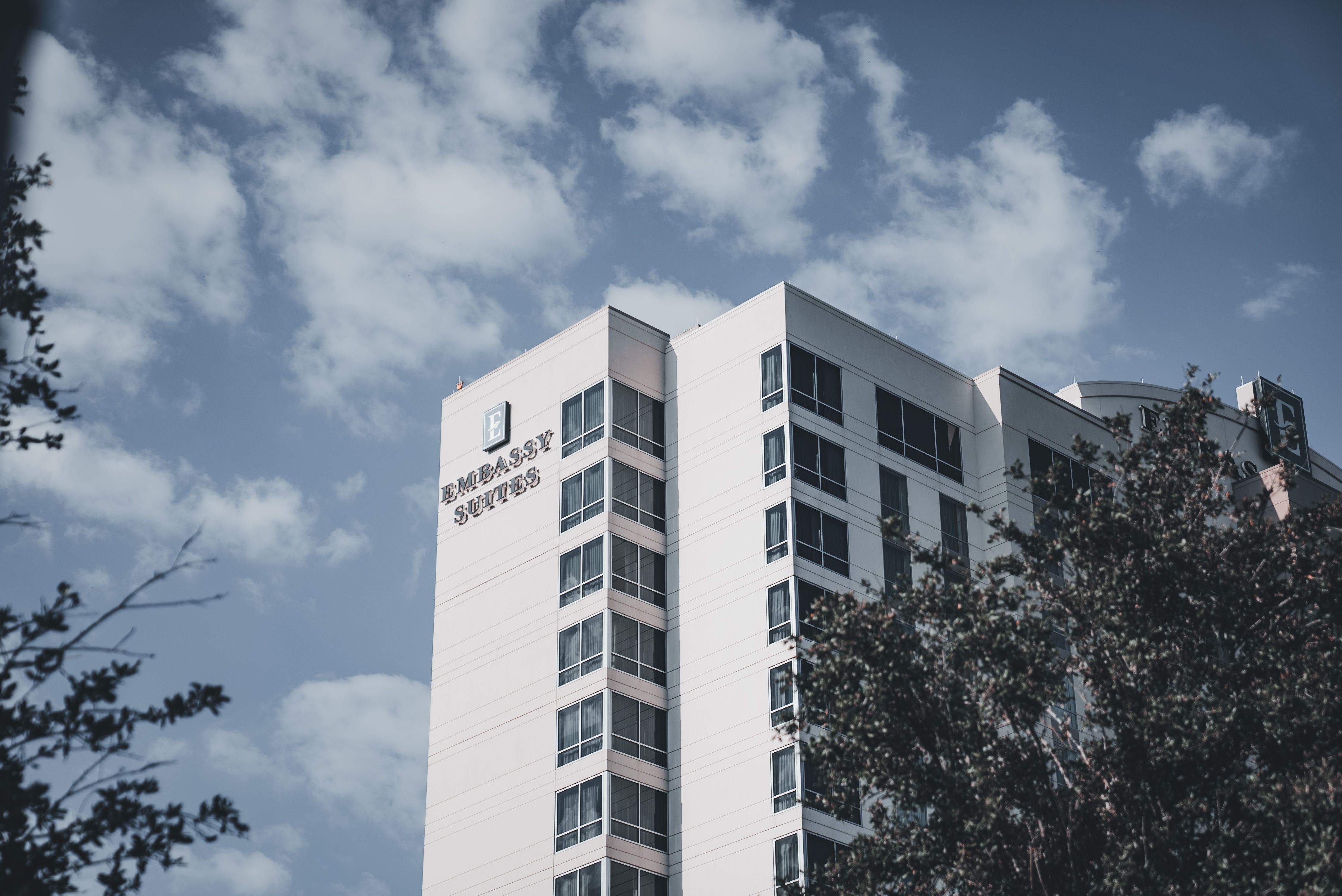 Elevated view of Embassy Suites hotel facade against a cloudy sky with trees in the foreground.