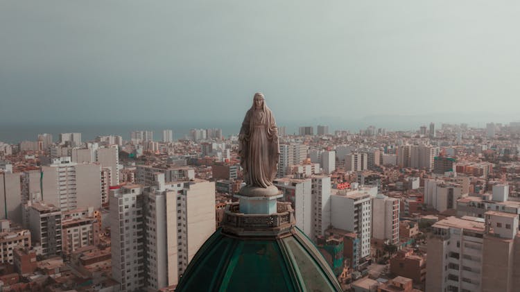 Virgin Mary Statue On Church Top Over City Buildings