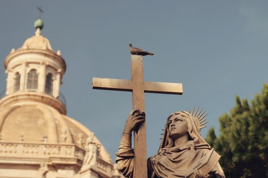 Statue holding a cross with a pigeon atop against a domed church in Catania, Sicily.