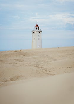 A lone lighthouse stands amidst vast sandy surroundings under a clear sky.