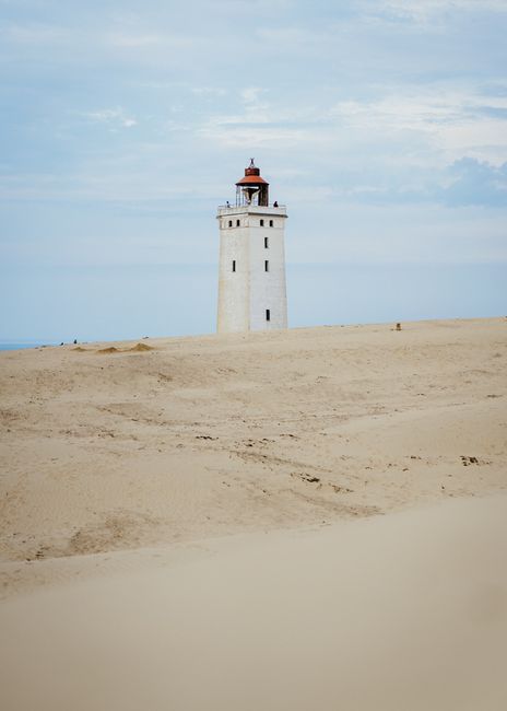 A lone lighthouse stands amidst vast sandy surroundings under a clear sky.
