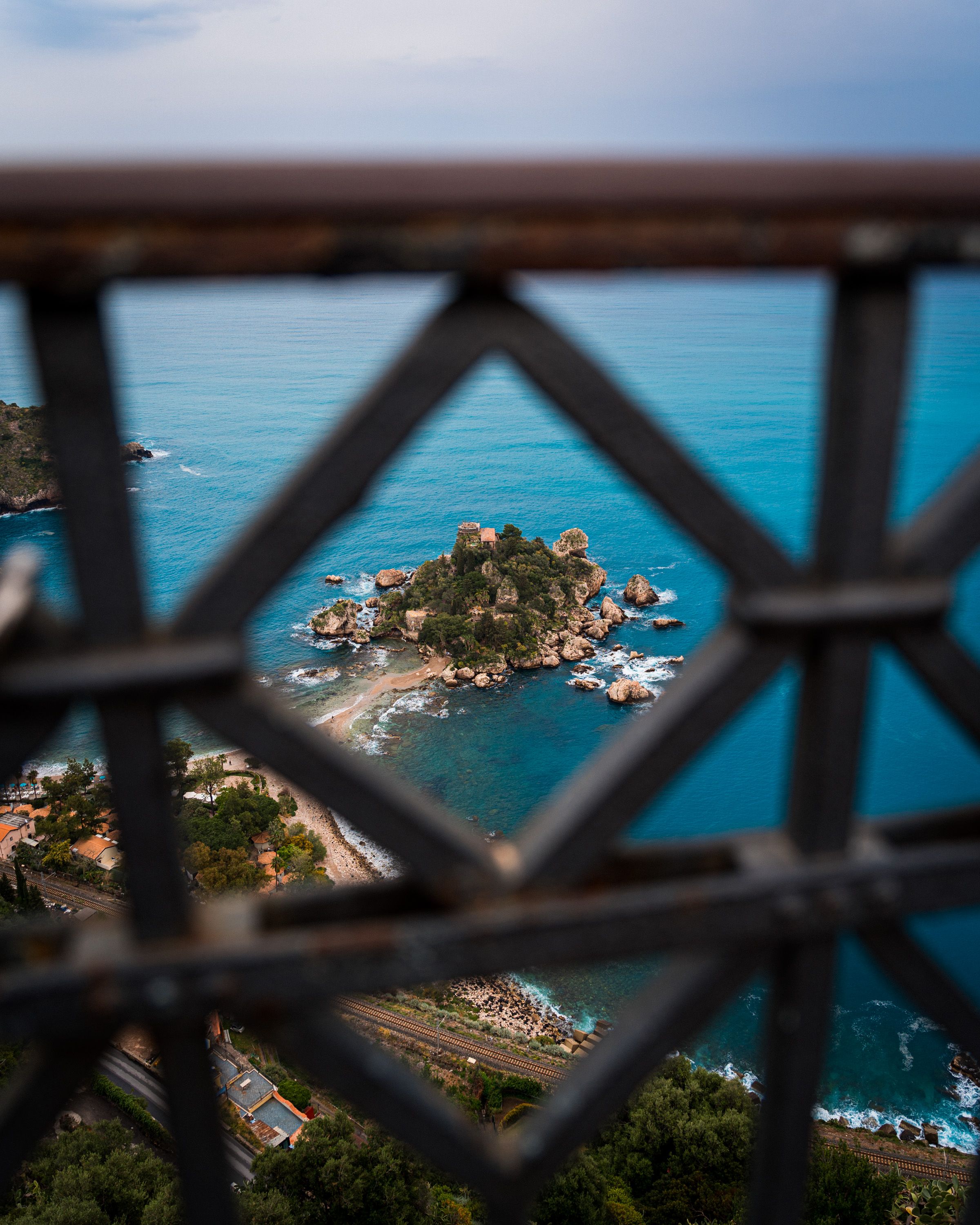 A picturesque coastal view in Sicily through decorative railings, showcasing a small rocky island and clear turquoise waters.