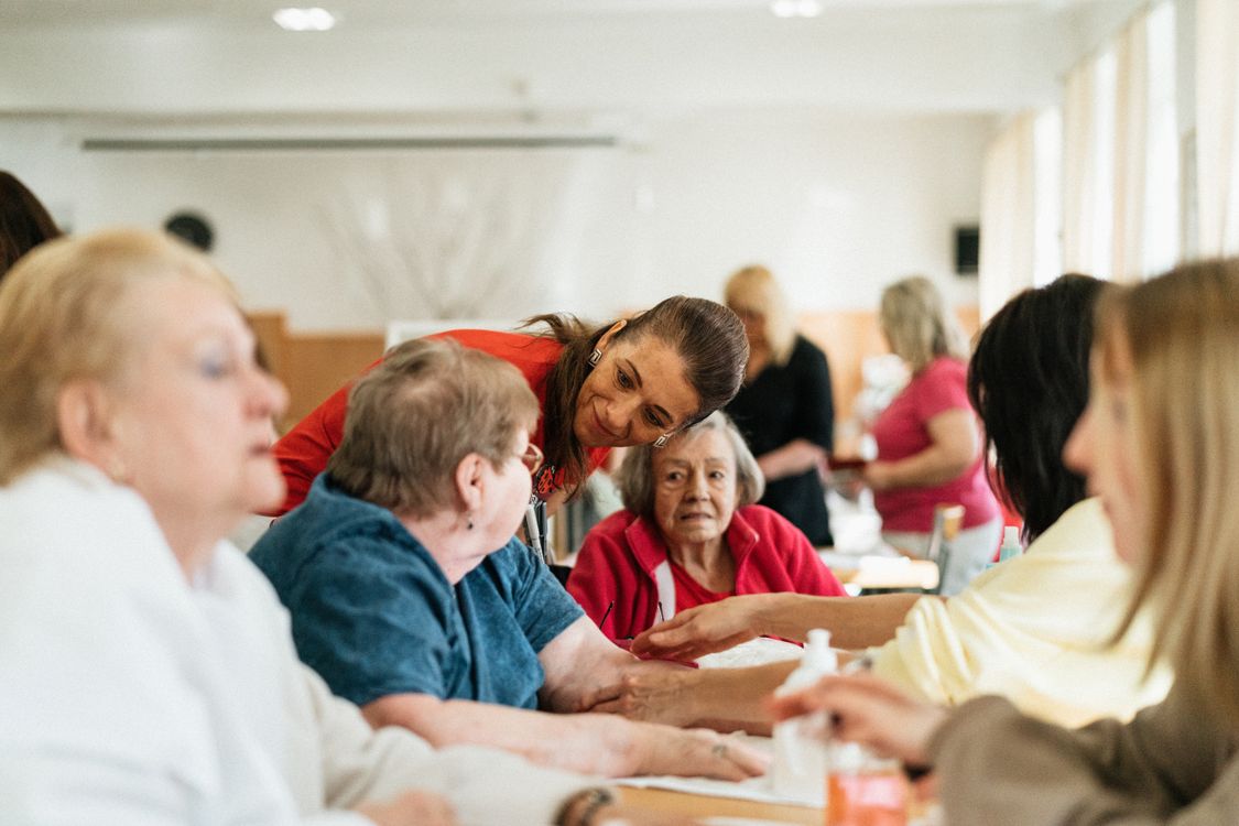 A caregiver smiling warmly while talking with a group of seniors seated at a table together. Photo by Jsme MILA on Pexels.
