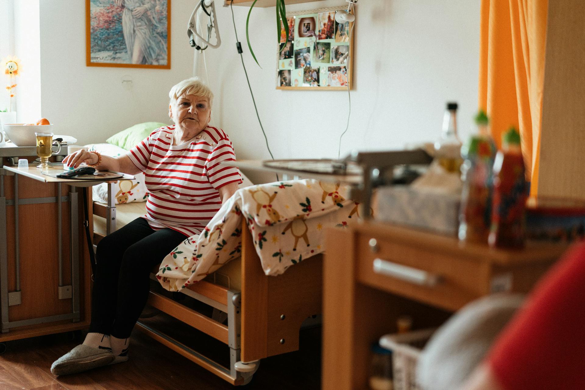 Elderly woman in striped shirt sits comfortably in her retirement home bedroom, surrounded by personal items.