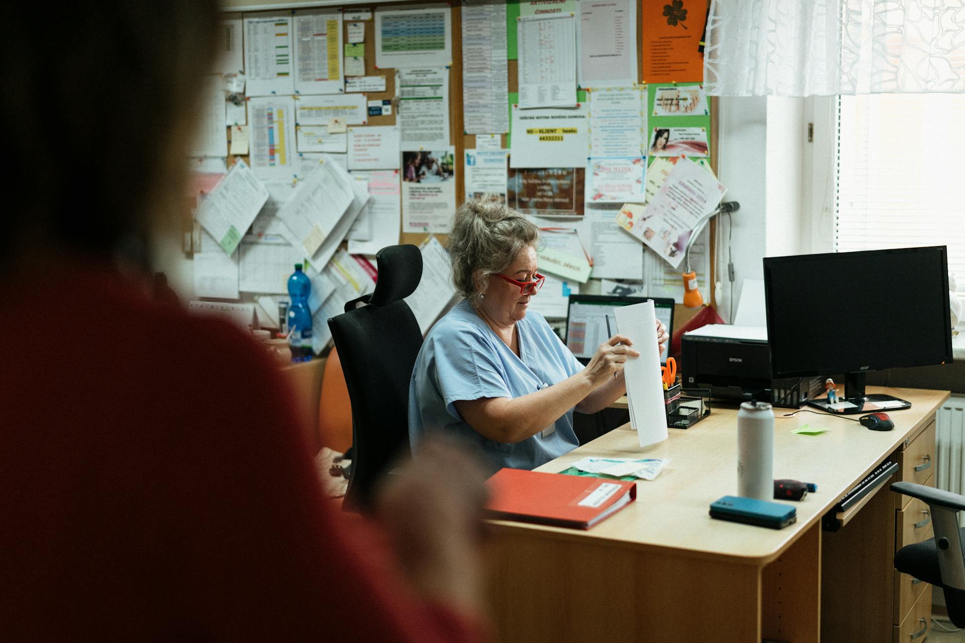 A senior caregiver in blue scrubs sorting paperwork at a hospital office desk.