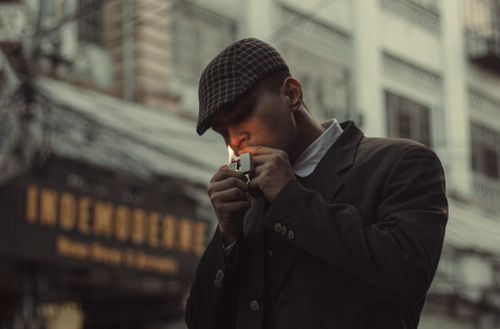 Free A man wearing a flat cap lights a cigarette in a city environment, adding moody urban charm. Stock Photo