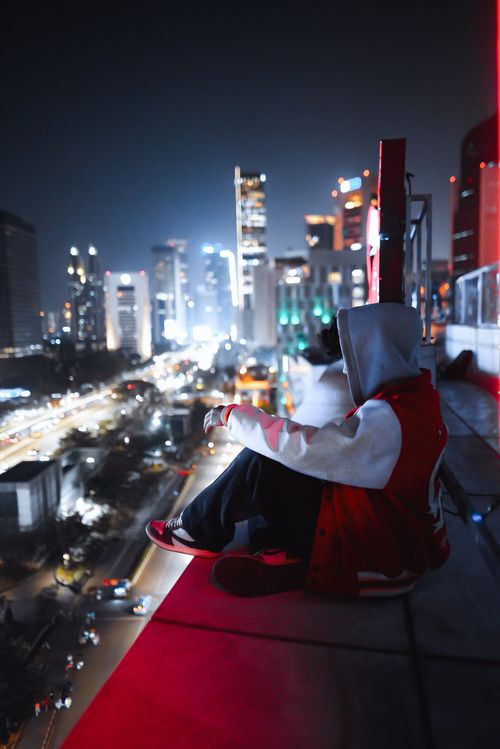 Free A person in a hoodie sits on a ledge, overlooking a brightly lit cityscape at night. Stock Photo