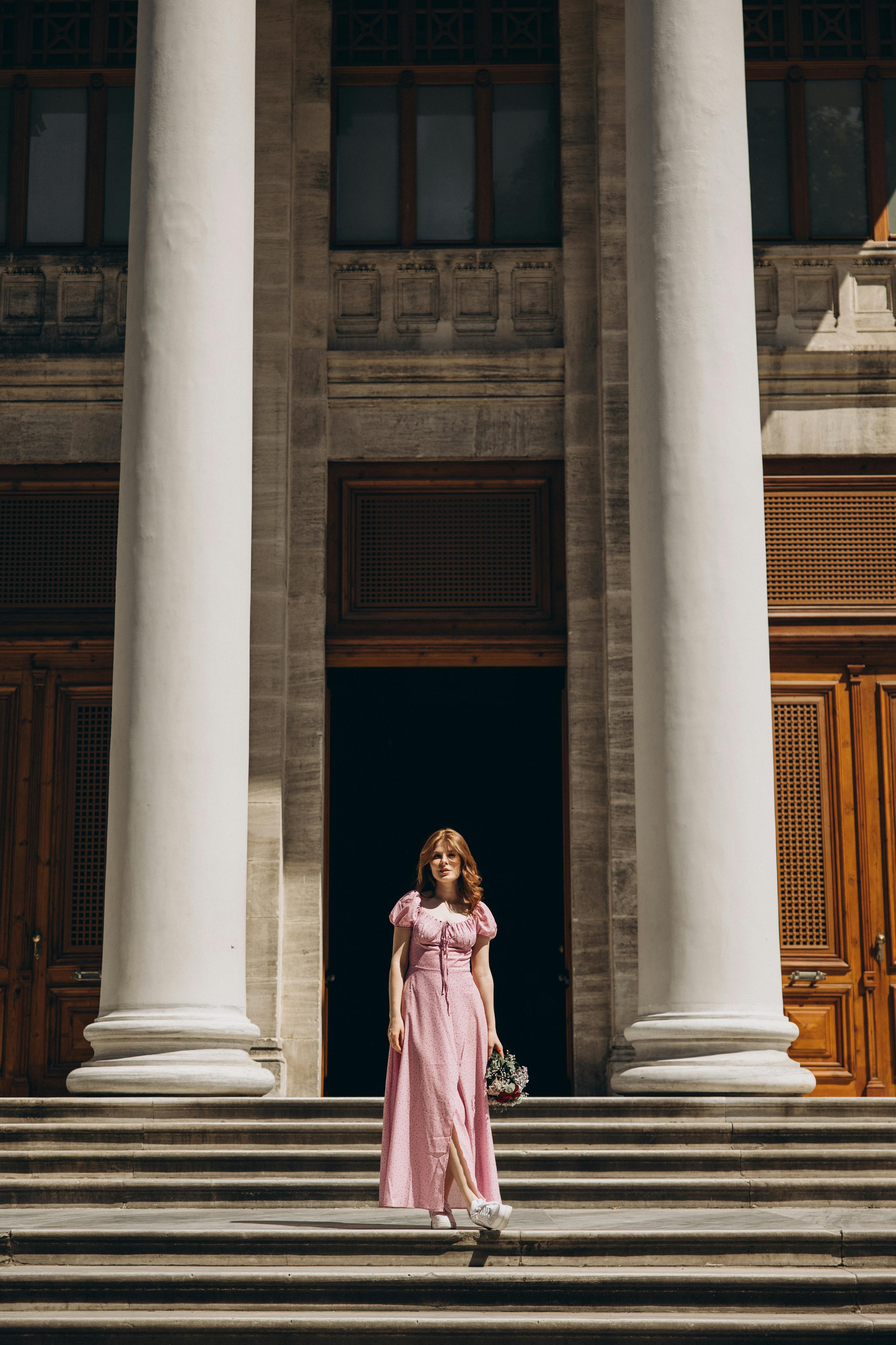 a woman in a pink dress stands on the steps of a building