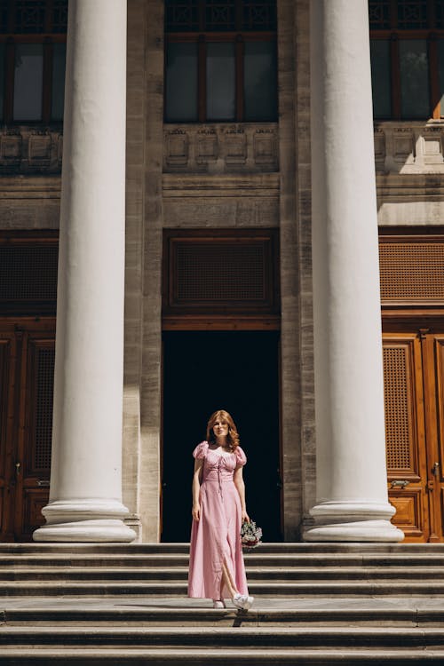 A woman in a pink dress stands on the steps of a building