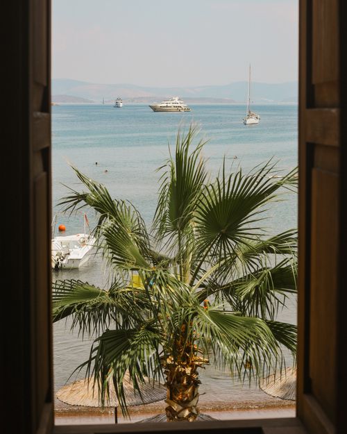 Free A serene view of boats on the tropical ocean framed by palm trees and wooden shutters. Stock Photo