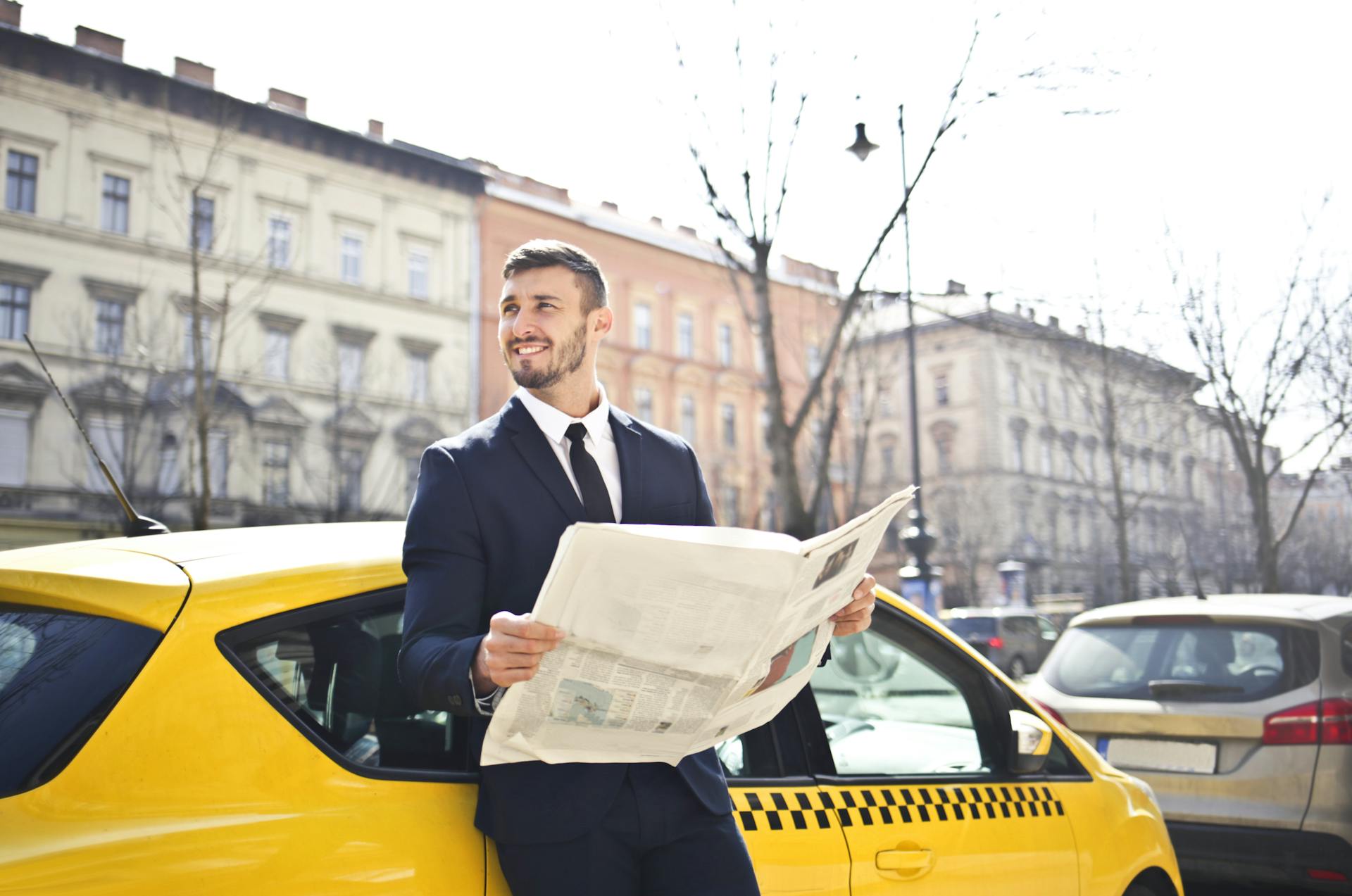 A businessman in a suit reads a newspaper while leaning on a yellow taxi in a city street.