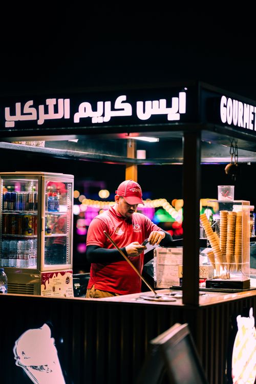 Free A man preparing ice cream cones at a nighttime illuminated booth with colorful lights. Stock Photo