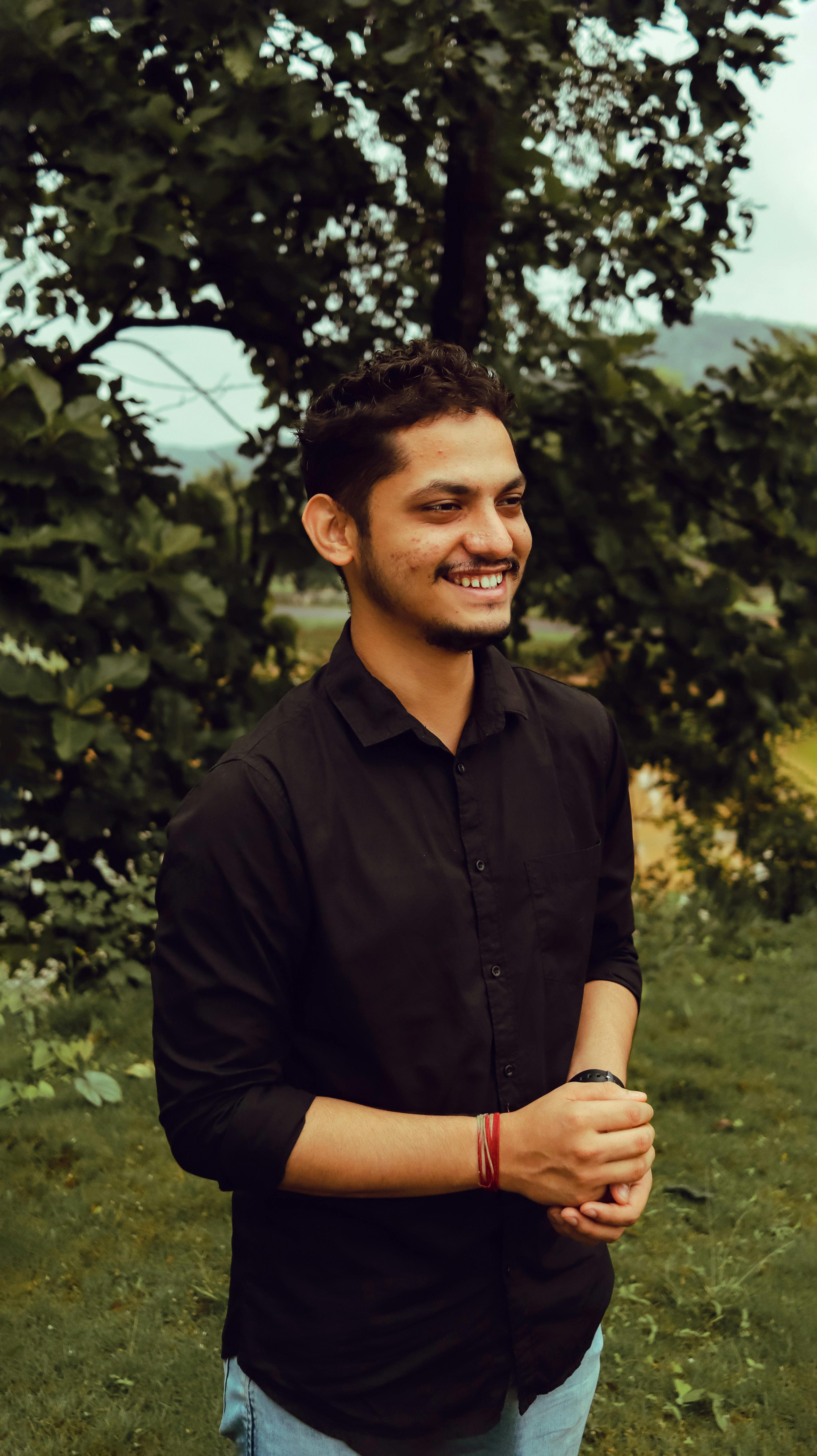 A cheerful young man in a black shirt smiling in an outdoor setting with lush greenery.