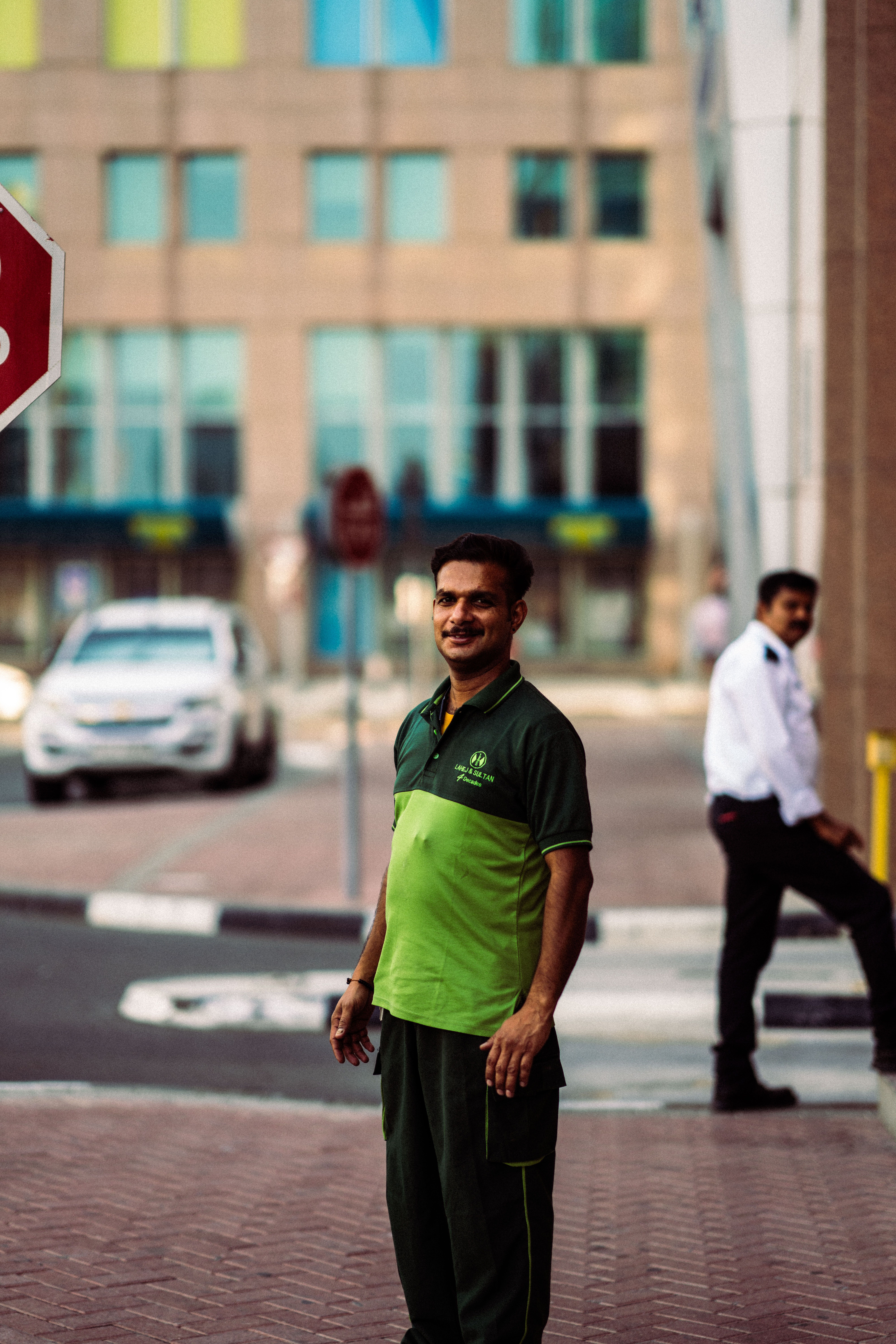 A smiling man stands on the sidewalk in an urban street scene, capturing the lively atmosphere.