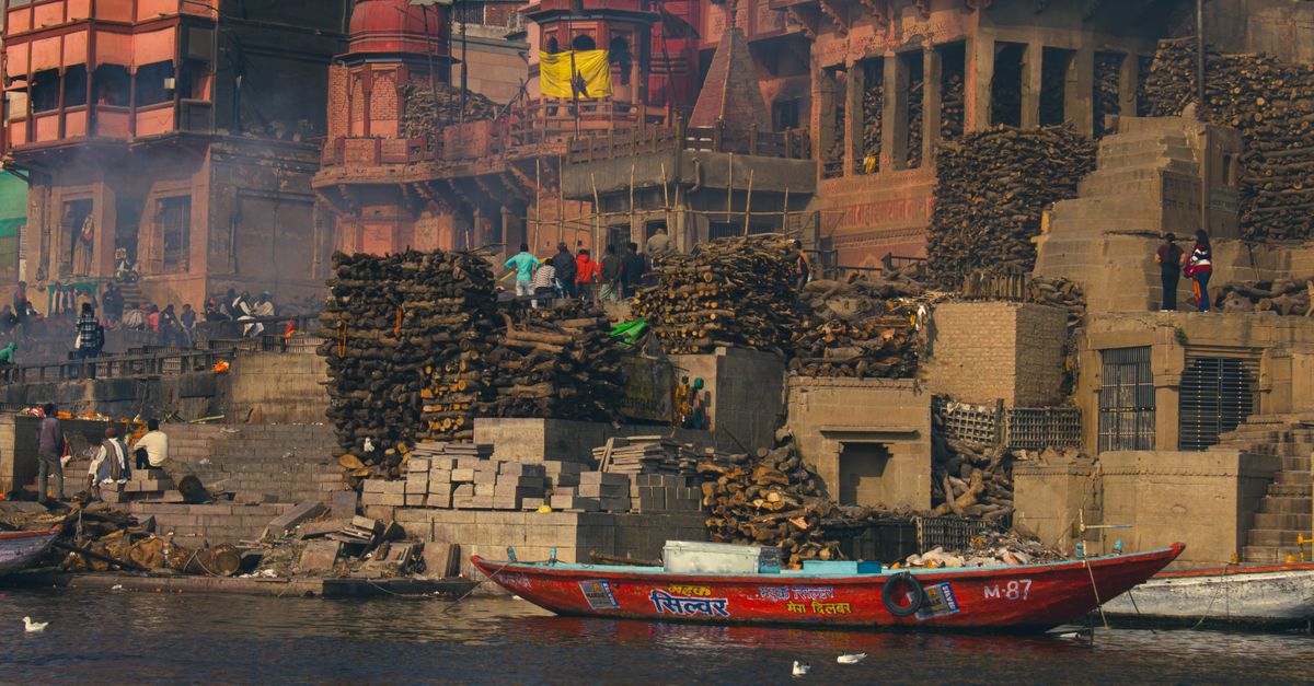 Colorful boats docked by ancient ghats with bustling urban scenery in the background.