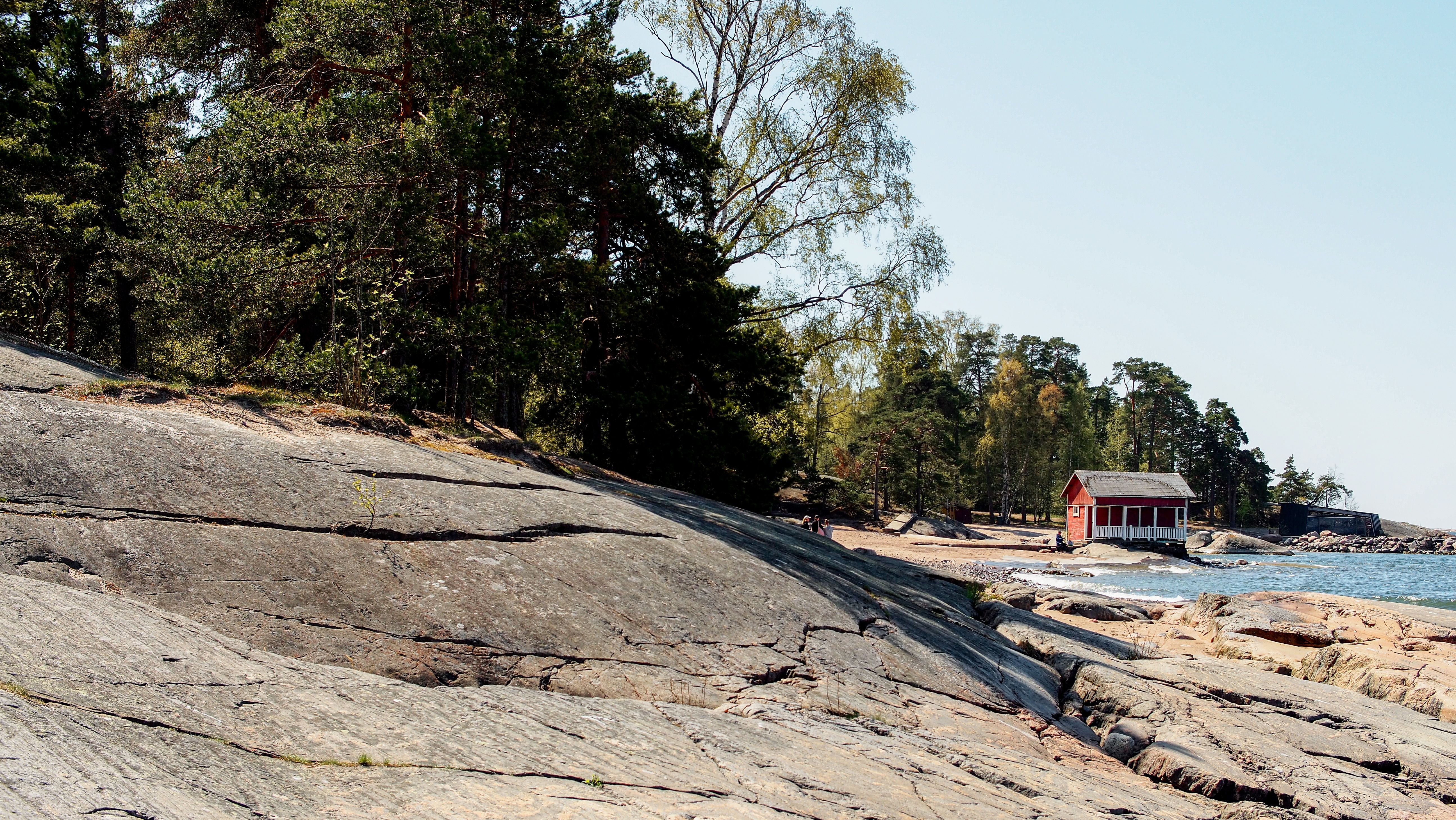 Natural coastal scenery in Helsinki with rocky shores and a red cabin, surrounded by lush trees.
