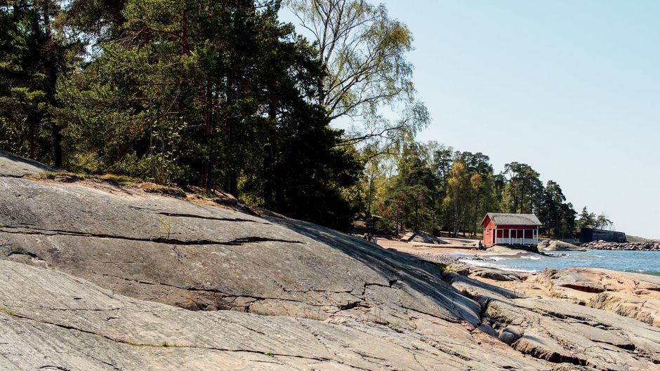 Natural coastal scenery in Helsinki with rocky shores and a red cabin, surrounded by lush trees.