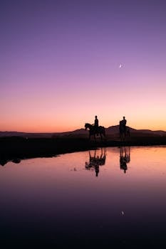 Two riders on horseback silhouetted against a purple twilight sky by a serene lake.