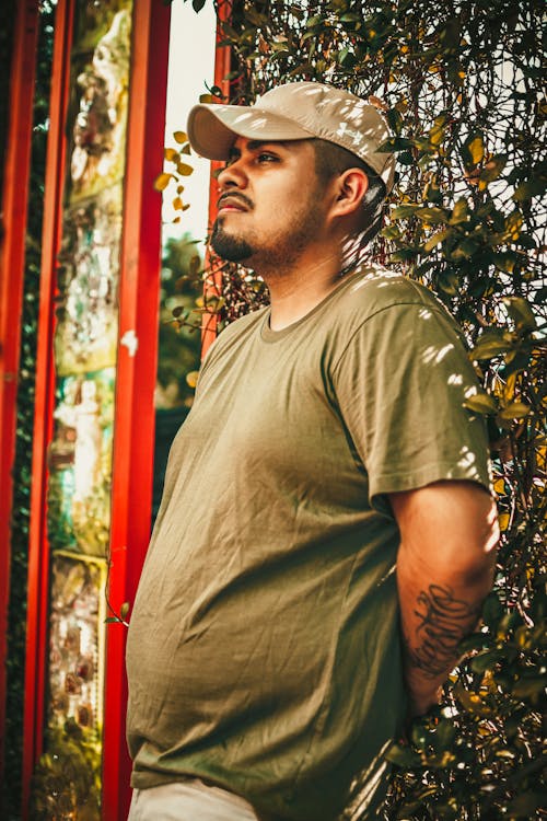 Man in a T-shirt and Cap Standing near a Hedge 